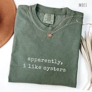 May include: A moss green t-shirt with the text "apparently, i like oysters" in white. A gold necklace with a pendant is draped over the shirt. The shirt is folded and has a tag that says "Comfort Colors".