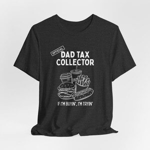 Może przedstawiać: Ciemnoszara koszulka z białym nadrukiem przedstawiającym jedzenie typu fast food, w tym burgera, frytki i hot doga. Tekst brzmi "DAD TAX COLLECTOR" i "IF I'M BUYIN', I'M TRYIN'" w kolorze białym.