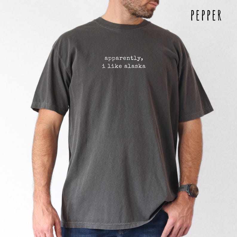 May include: Pepper-colored t-shirt with the text "apparently, i like alaska" in white. The shirt is a dark gray color and has short sleeves. The text is centered on the chest.
