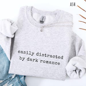 May include: A light gray sweatshirt with the text "easily distracted by dark romance" in black. A silver necklace with a small pendant rests on the sweatshirt. The sleeves are cuffed, and a portion of blue jeans is visible.