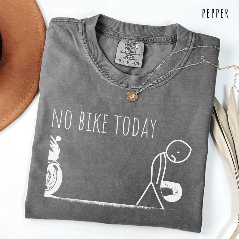 May include: Pepper-colored Comfort Colors t-shirt with the text "NO BIKE TODAY" and a graphic of a motorcycle and a dejected person. A silver necklace with a pendant is draped across the shirt.
