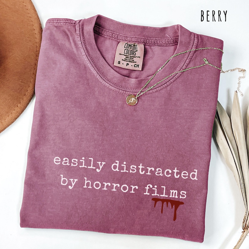 May include: Berry-colored Comfort Colors t-shirt with the text "easily distracted by horror films" in white. A gold necklace with a pendant rests on the shirt. The shirt has a small blood drip graphic.