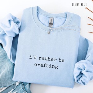 Pu&ograve; includere: Felpa azzurra con la scritta nera "i'd rather be crafting". Una collana d'argento con un ciondolo a forma di fiore &egrave; sulla felpa. La felpa &egrave; piegata sopra un paio di jeans blu.