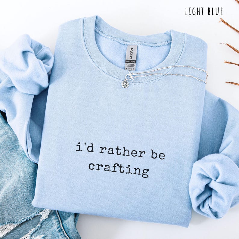 Pu&ograve; includere: Felpa azzurra con la scritta nera "i'd rather be crafting". Una collana d'argento con un ciondolo a forma di fiore &egrave; sulla felpa. La felpa &egrave; piegata sopra un paio di jeans blu.