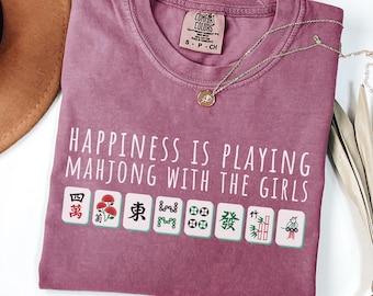 May include: A berry-colored short-sleeve t-shirt featuring the white text "HAPPINESS IS PLAYING MAHJONG WITH THE GIRLS" above a row of eight colorful mahjong tiles. The shirt has a crew neckline and a Comfort Colors brand label at the inner collar.