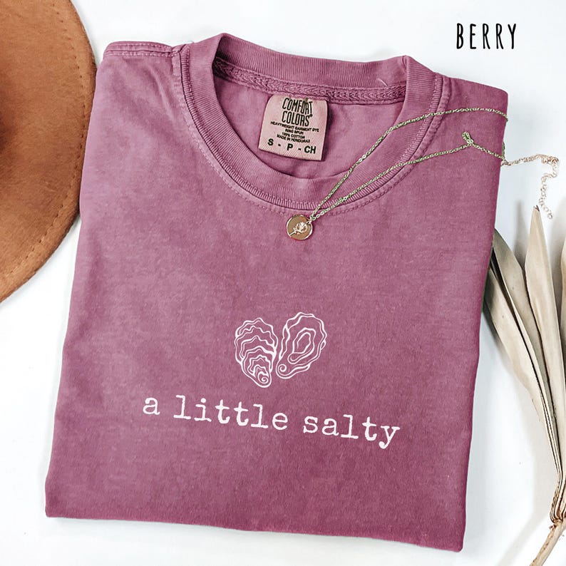 Pode incluir: Camiseta cor de berry com o texto "a little salty" e um gr&aacute;fico de duas conchas de ostra. Um colar de ouro com um pingente repousa sobre a camisa. A camisa &eacute; da marca Comfort Colors.