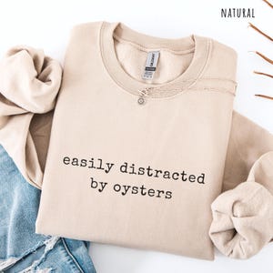 May include: A beige sweatshirt with the text "easily distracted by oysters" in black. A silver necklace with a sun pendant is on the sweatshirt. The sweatshirt is folded with a pair of blue jeans and a beige sleeve.