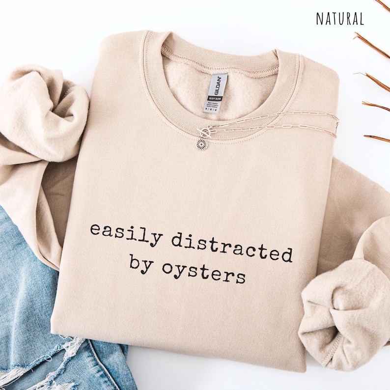 May include: A beige sweatshirt with the text "easily distracted by oysters" in black. A silver necklace with a sun pendant is on the sweatshirt. The sweatshirt is folded with a pair of blue jeans and a beige sleeve.