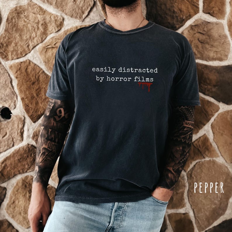 May include: Dark gray t-shirt with the text "easily distracted by horror films" in white, with a blood-like drip effect. The shirt is worn by a person with tattoos on their arms and is paired with blue jeans.