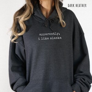 May include: Dark heather gray hoodie with the text "apparently, i like alaska" in white. The hoodie has a drawstring hood and a front pocket. The words are in a simple, sans-serif font.