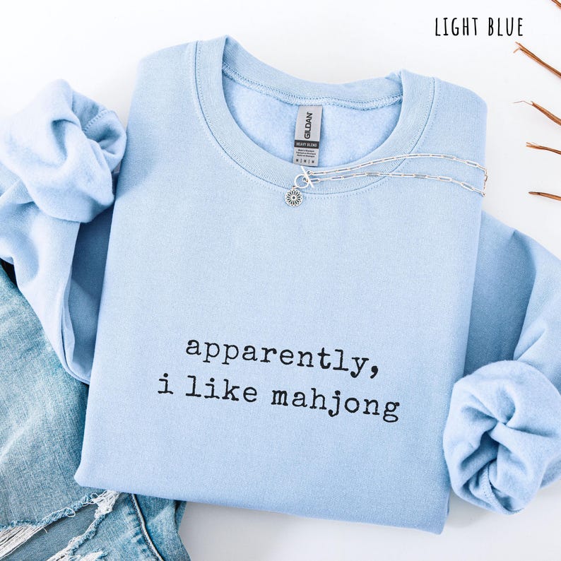 May include: Light blue sweatshirt with the text "apparently, i like mahjong" in black. A silver necklace with a small pendant is on the shirt. The sweatshirt is folded with a pair of blue jeans.