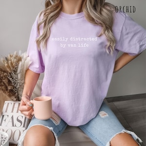 May include: A light purple t-shirt with the text "easily distracted by van life" in white. The shirt is worn with light blue jeans. A peach-colored mug is held in the hand. The word "ORCHID" is printed in the upper right corner.
