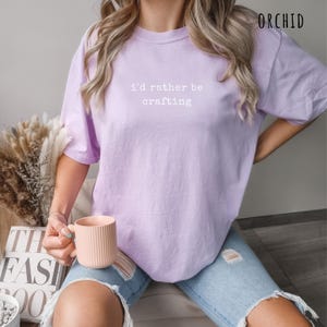 Pu&ograve; includere: Una maglietta lilla con la scritta bianca "I'd rather be crafting". La maglietta &egrave; realizzata in un materiale morbido e confortevole. La parola "ORCHID" &egrave; stampata nell'angolo in alto a destra.