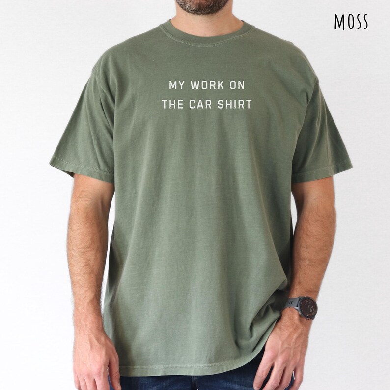 May include: A moss green t-shirt with the text "MY WORK ON THE CAR SHIRT" in white. The shirt is a crew neck style and has short sleeves. The word "MOSS" is printed in the upper right corner.