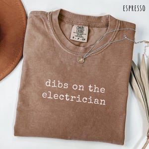 May include: Espresso-colored t-shirt with the phrase "dibs on the electrician" in white lettering. A silver necklace with a small pendant rests on the shirt. The shirt is a Comfort Colors brand, size S-CH.
