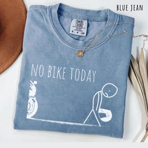 May include: Blue jean Comfort Colors t-shirt with the text "NO BIKE TODAY" and a graphic of a motorcycle and a dejected stick figure. A gold necklace with a pendant is draped across the shirt.