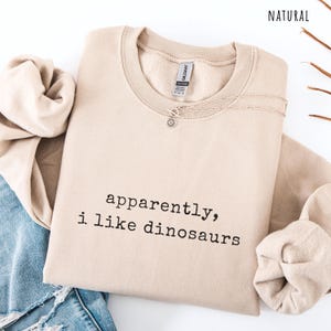 May include: A beige sweatshirt with the text "apparently, i like dinosaurs" in black. A silver necklace with a sun pendant is on the sweatshirt. The word "NATURAL" is in the top right corner.