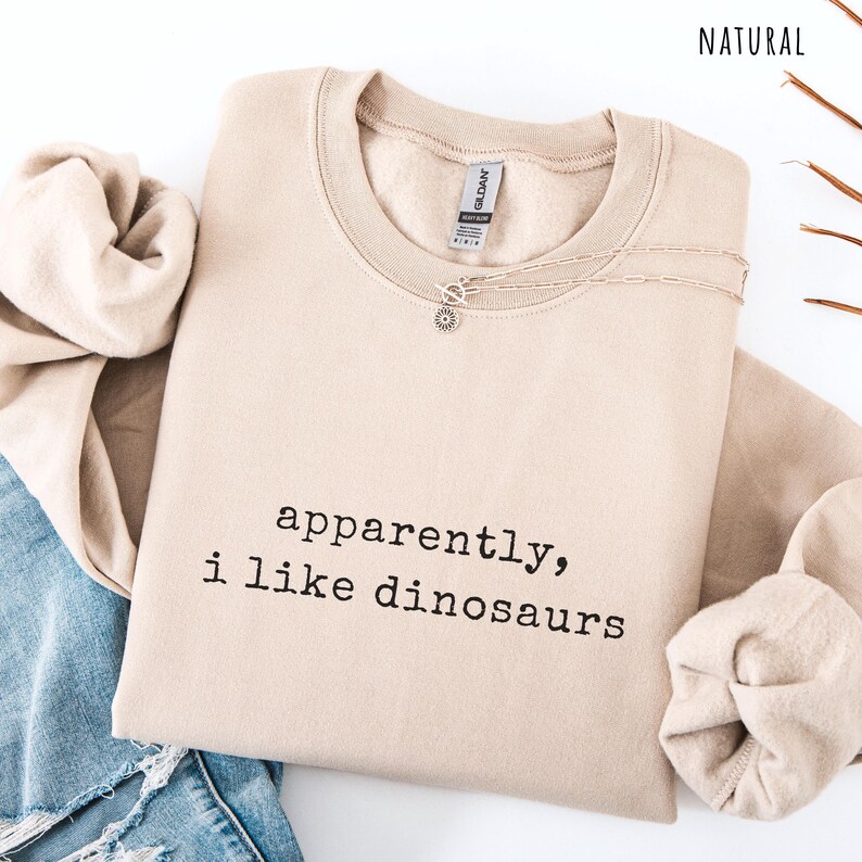 May include: A beige sweatshirt with the text "apparently, i like dinosaurs" in black. A silver necklace with a sun pendant is on the sweatshirt. The word "NATURAL" is in the top right corner.