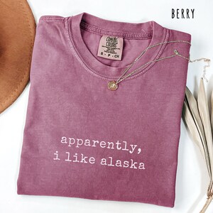 May include: Berry-colored t-shirt with the text "apparently, i like alaska" in white. The shirt has a crew neck and short sleeves. A gold necklace with a pendant is draped across the shirt.