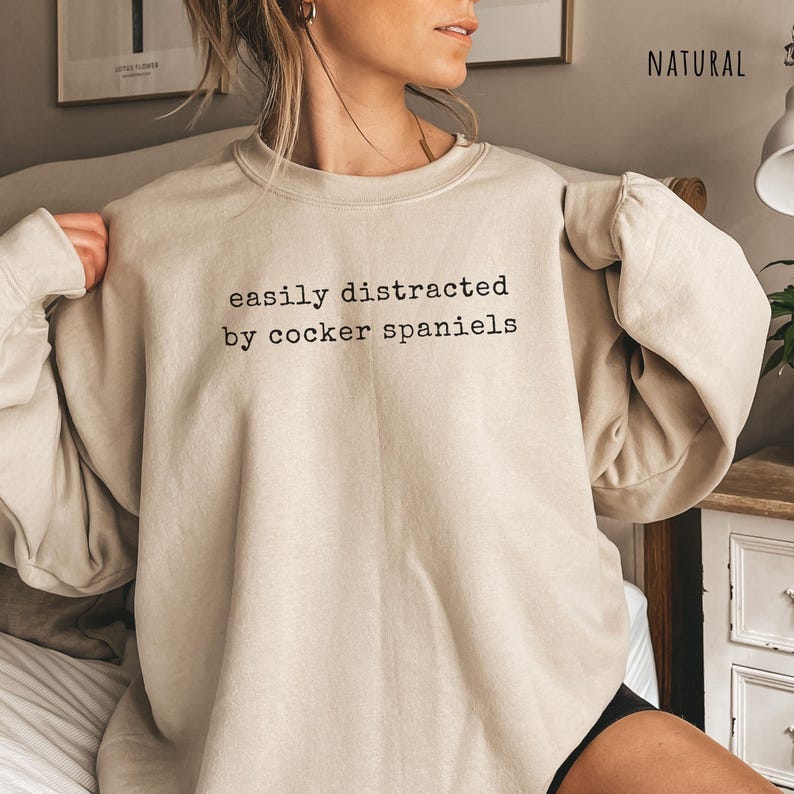 May include: A beige sweatshirt with the text "easily distracted by cocker spaniels" in black. The long-sleeved crew neck top is made of a soft material and is a neutral color.
