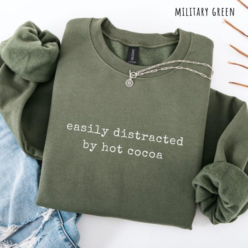 May include: A military green crewneck sweatshirt featuring the white text "easily distracted by hot cocoa" printed on the chest. The garment has long sleeves with ribbed cuffs and a ribbed neckline. A silver-toned chain necklace rests on the collar.