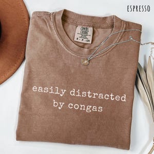 May include: An espresso-colored short-sleeve t-shirt with a crew neckline. The shirt features the white text 'easily distracted by congas' printed on the front. A silver-toned necklace with a small circular pendant rests on the shirt collar.