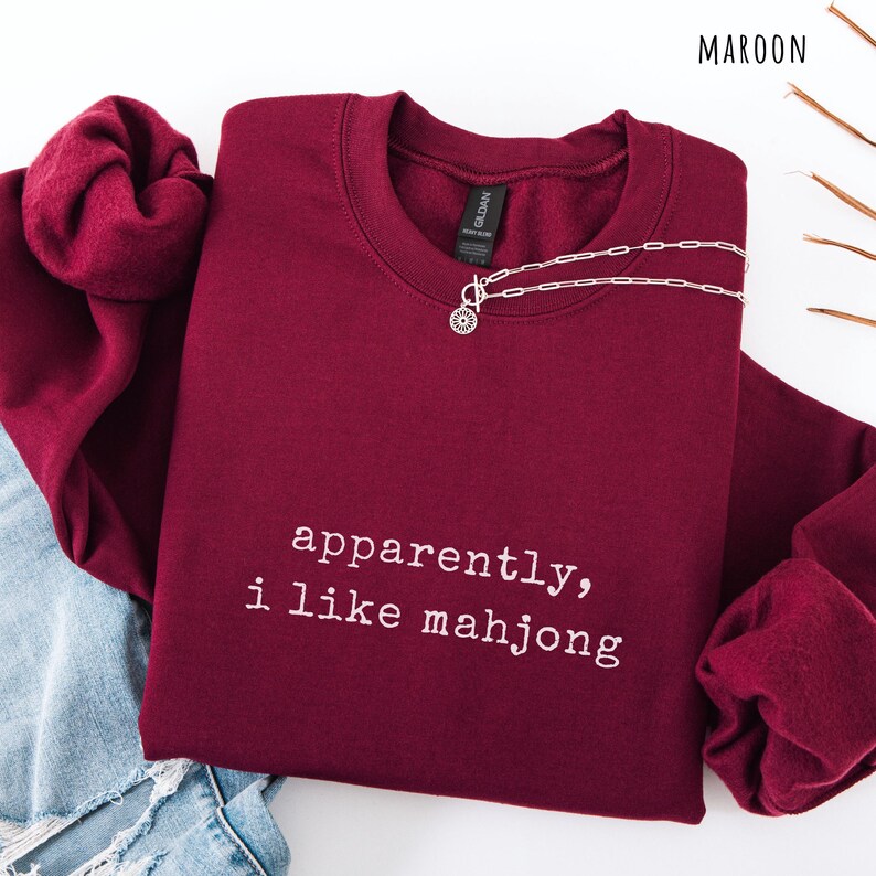 May include: Maroon crewneck sweatshirt with the text "apparently, i like mahjong" in white. A silver necklace with a circular pendant is draped on the sweatshirt. The word "MAROON" is in the top right corner.