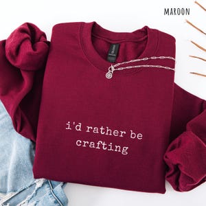 Pu&ograve; includere: Una felpa girocollo bordeaux con la scritta bianca "i'd rather be crafting". Una collana d'argento con un ciondolo circolare &egrave; drappeggiata sul davanti. La parola "MAROON" &egrave; nell'angolo in alto a destra.