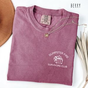 May include: A berry-colored t-shirt with the text "Dumpster Fire Survivors Club" in white. The shirt has a crew neck and short sleeves. A gold necklace with a pendant is draped across the shirt.