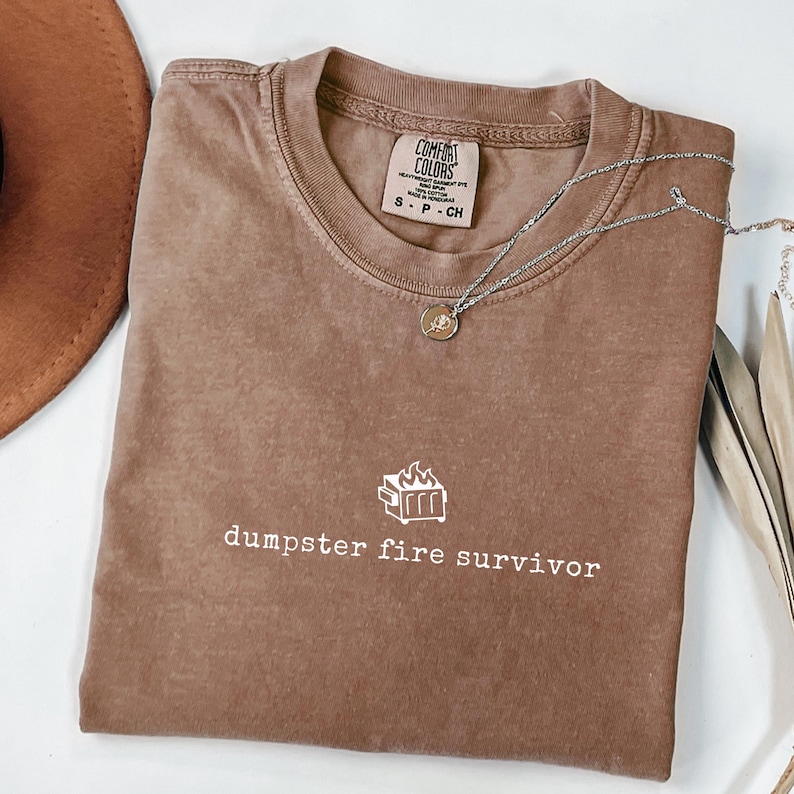 May include: A brown t-shirt with the text "dumpster fire survivor" and a small graphic of a dumpster with flames on it.