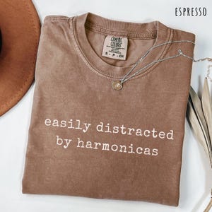 May include: An espresso-colored t-shirt with a crew neckline and a Comfort Colors brand label. The shirt features the white text "easily distracted by harmonicas" printed on the front. A silver-toned chain necklace with a small circular pendant rests on the shirt.
