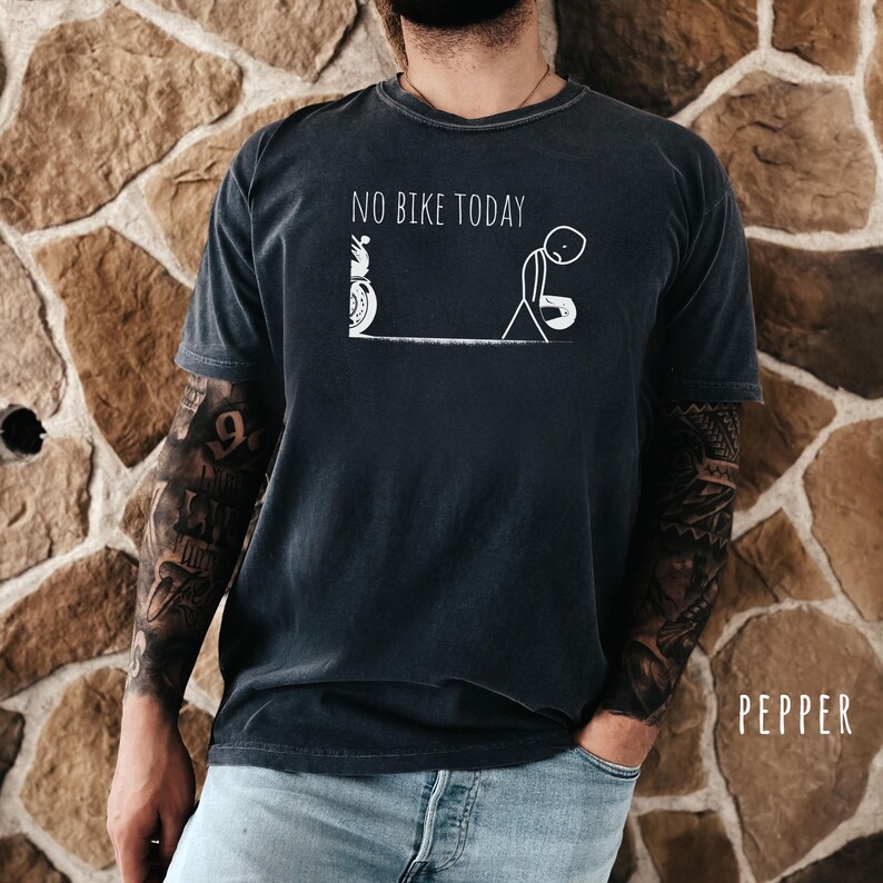 May include: Dark gray t-shirt with the text "NO BIKE TODAY" and a stick figure design of a bicycle and a person. The shirt is worn by a person with tattoos on their arms and is paired with blue jeans.