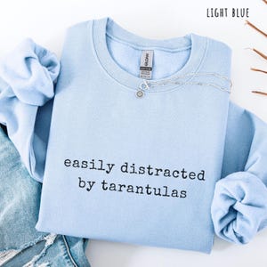 May include: Light blue sweatshirt with the text "easily distracted by tarantulas" in black. A silver necklace with a flower pendant is on the shirt. The sweatshirt is folded on top of a pair of blue jeans.