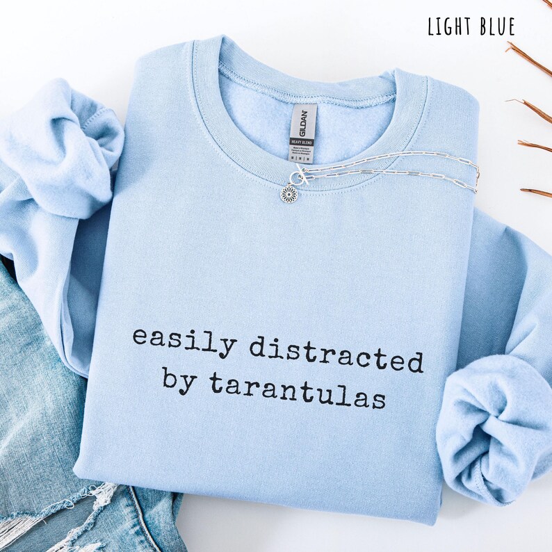 May include: Light blue sweatshirt with the text "easily distracted by tarantulas" in black. A silver necklace with a flower pendant is on the shirt. The sweatshirt is folded on top of a pair of blue jeans.