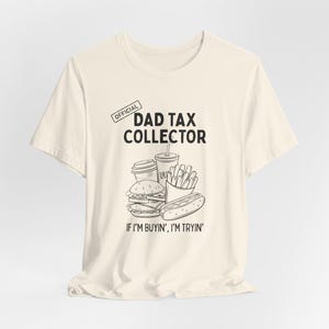 Może przedstawiać: Kremowa koszulka z napisem "OFFICIAL DAD TAX COLLECTOR" i grafiką fast food. Poniżej grafiki znajduje się fraza "IF I'M BUYIN', I'M TRYIN'".