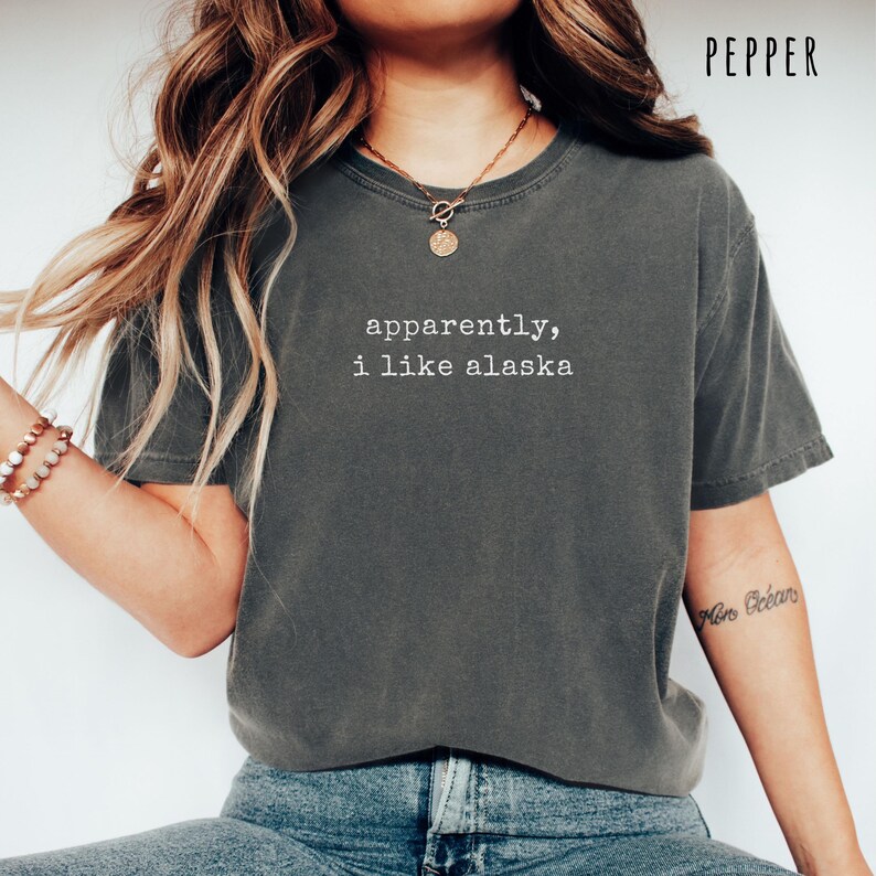 May include: Pepper-colored t-shirt with the text "apparently, i like alaska" in white. The shirt is a relaxed fit, with short sleeves and a crew neck. The shirt is made of a soft, comfortable material.