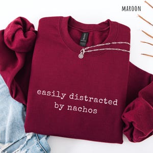 May include: Maroon sweatshirt with the phrase "easily distracted by nachos" in white lettering. A silver necklace with a pendant is draped on the sweatshirt. The sweatshirt is folded on top of a pair of blue jeans.