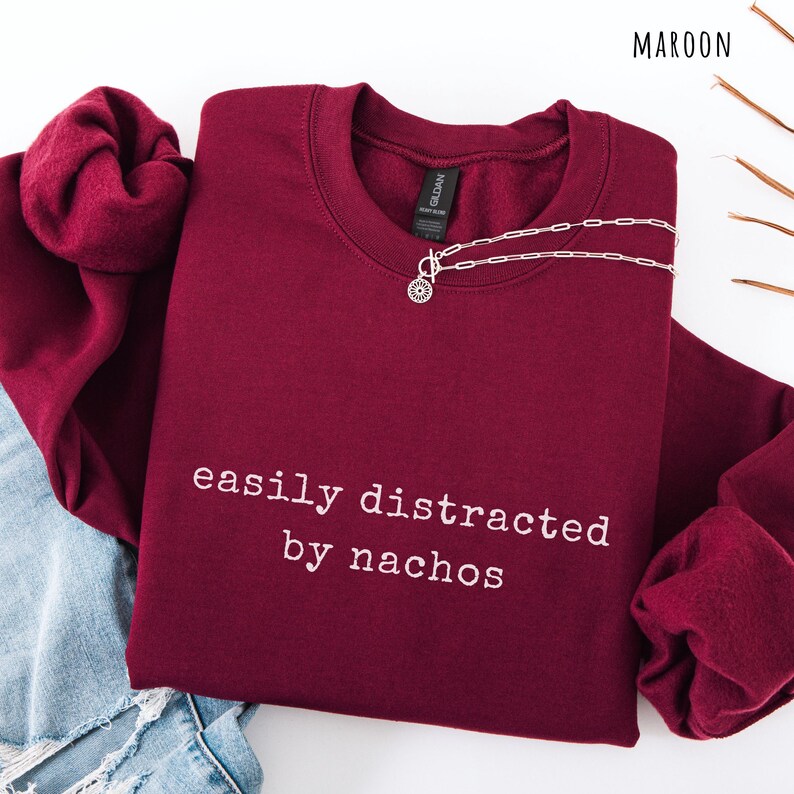 May include: Maroon sweatshirt with the phrase "easily distracted by nachos" in white lettering. A silver necklace with a pendant is draped on the sweatshirt. The sweatshirt is folded on top of a pair of blue jeans.