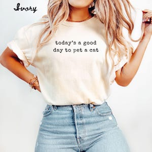 Op de afbeelding: Ivoor kleurig t-shirt met de zwarte tekst "today's a good day to pet a cat". Het shirt heeft opgerolde mouwen en wordt gecombineerd met een blauwe jeans. Het woord "Ivory" staat in cursief linksboven.