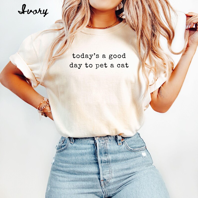 Op de afbeelding: Ivoor kleurig t-shirt met de zwarte tekst "today's a good day to pet a cat". Het shirt heeft opgerolde mouwen en wordt gecombineerd met een blauwe jeans. Het woord "Ivory" staat in cursief linksboven.