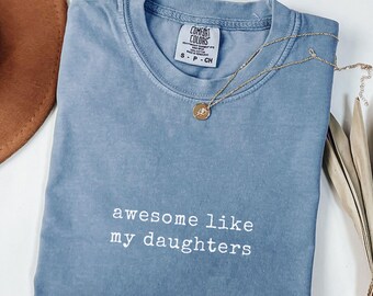May include: A blue jean colored Comfort Colors t-shirt with the text "awesome like my daughters" in white. A gold necklace with a pendant is draped across the shirt. The shirt is folded and ready to wear.