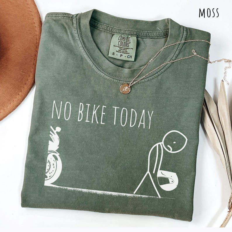 May include: Moss green t-shirt with the text "NO BIKE TODAY" and a graphic of a person and a motorcycle. A gold necklace with a pendant is on the shirt. The shirt is folded and has a tag that says "Comfort Colors".