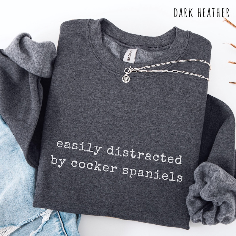 May include: Dark heather gray sweatshirt with the text "easily distracted by cocker spaniels" in white. A silver necklace with a sunburst pendant is draped across the top. The sweatshirt is folded with the sleeves visible.