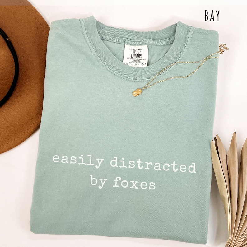 Pu&ograve; includere: T-shirt verde chiaro con la scritta bianca "easily distracted by foxes". La maglietta ha girocollo e maniche corte. Una collana d'oro con un ciondolo &egrave; drappeggiata sulla maglietta.