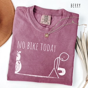 May include: Berry-colored t-shirt with the text "NO BIKE TODAY" and a graphic of a motorcycle and a person. The shirt has a crew neck and short sleeves. A gold necklace with a pendant is draped across the shirt.