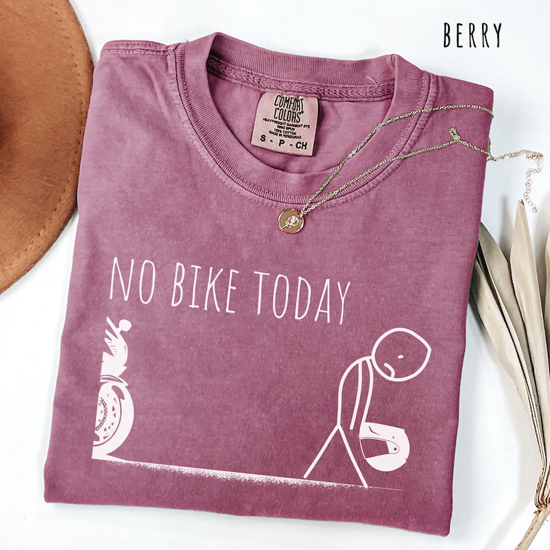 May include: Berry-colored t-shirt with the text "NO BIKE TODAY" and a graphic of a motorcycle and a person. The shirt has a crew neck and short sleeves. A gold necklace with a pendant is draped across the shirt.