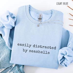 Peut inclure: Sweat-shirt bleu clair avec l'inscription noire "easily distracted by seashells". Un collier argent&eacute; avec un pendentif en forme de fleur est pos&eacute; sur le sweat-shirt. L'inscription "light blue" est en haut &agrave; droite.