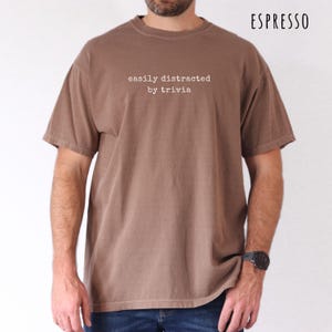 May include: Espresso-colored t-shirt with the text "easily distracted by trivia" in a white, sans-serif font. The shirt has a relaxed fit and short sleeves. The word "ESPRESSO" is printed in the upper right corner.