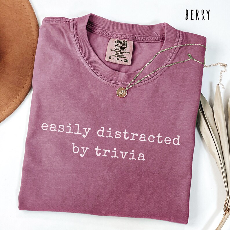 May include: Berry-colored t-shirt with the phrase "easily distracted by trivia" in white. A gold-colored necklace with a pendant is draped across the shirt. The shirt has a crew neck and a Comfort Colors tag.