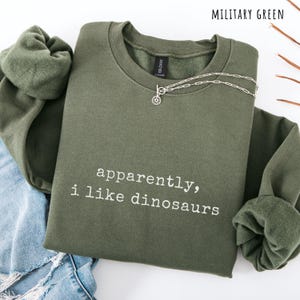 May include: Military green sweatshirt with the text "apparently, i like dinosaurs" in white. A silver necklace with a sunburst pendant is draped over the front. The sweatshirt is folded on top of a pair of blue jeans.